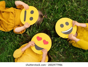 Three children lying on the grass are holding cardboard emoticons with different emotions in their hands: a sad, smiling happy smile, a loving smile with hearts instead of eyes.  World Emoji Day - Shutterstock ID 2002977986