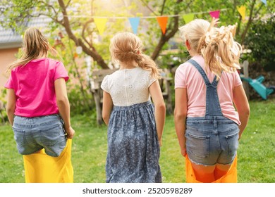 Three children are outdoors in a grassy garden, participating in a fun sack race with colorful bags. Bunting flags hang in the background, creating a festive and playful atmosphere. - Powered by Shutterstock