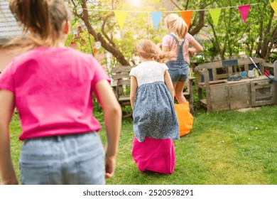 Three children are outdoors in a grassy garden, participating in a fun sack race with colorful bags. Bunting flags hang in the background, creating a festive and playful atmosphere. - Powered by Shutterstock