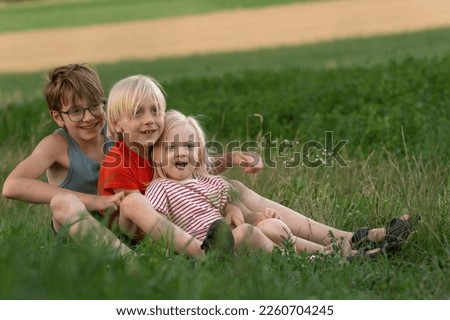 Three children sit on green grass and smile. Portrait of siblings outside. Two brothers and little sister are walking outdoors