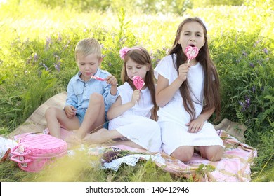 Three children sitting on the rug in the grass and eat lollipops outdoors
