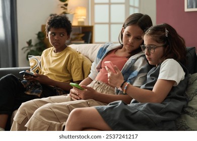 Three children sitting on sofa playing mobile games together and sharing in their excitement for technology. Young minds are engaging with handheld devices in cozy indoor setting - Powered by Shutterstock