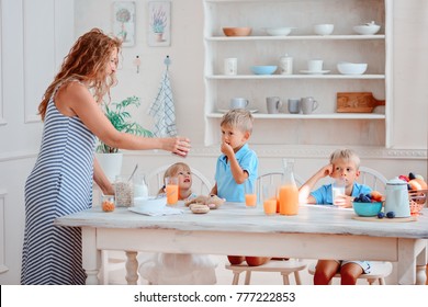 Three Children Are Sitting At The Table And Their Mother Is Feeding Them Breakfast / Big Family Is Having Breakfast At A Big Table, On The Table Are Juice, Oatmeal, Plates And Fruits, Berries