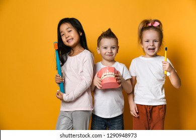 Three children with toothbrushes and a Dental implant model  stand over yellow background wall and smile rejoicing at the camera.The concept of health, oral hygiene, people and beauty. - Shutterstock ID 1290948973