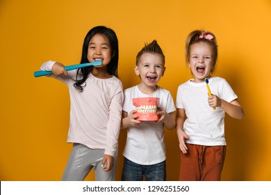 Three children with toothbrushes and a Dental implant model  stand over yellow background wall and smile rejoicing at the camera.The concept of health, oral hygiene, people and beauty.