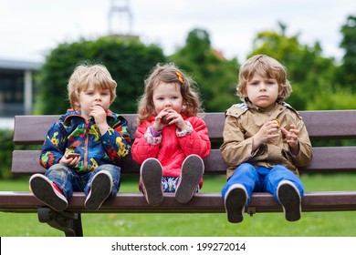 Three children: two little boys and one girl sitting on bench and eating chocolate