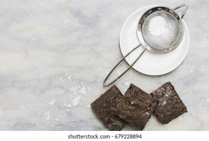 Three Chocolate Brownies With Icing Sugar In Sifter On Marble Background  From Overhead View.
