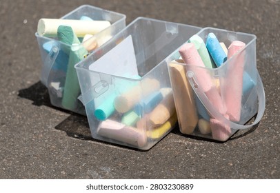 Three clear containers hold assorted chalks. Colors include green, pink, blue, and yellow. Chalk pieces stand upright in the bins. They rest on a dark asphalt surface