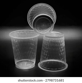 Three clear, disposable plastic drinking cups featuring a textured exterior pattern are arranged against a stark black background. The cup on the left stands upright on its flat base, showcasing a wide rim and a body covered in an embossed, diamond-shaped grid pattern that mimics quilted or faceted glass to improve grip and structural strength. The cup on the right is inverted, resting upside down on its rim to display the continuation of the textured design along its tapered sides and a smooth, untextured band near the bottom edge.Balancing precariously on top of the inverted cup is the third container, tilted directly toward the viewer to prominently reveal its underside. The flat, circular base of this top cup features raised, molded text and symbols, including a prominent number 