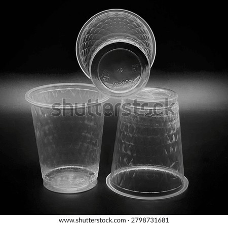 Three clear, disposable plastic drinking cups featuring a textured exterior pattern are arranged against a stark black background. The cup on the left stands upright on its flat base, showcasing a wide rim and a body covered in an embossed, diamond-shaped grid pattern that mimics quilted or faceted glass to improve grip and structural strength. The cup on the right is inverted, resting upside down on its rim to display the continuation of the textured design along its tapered sides and a smooth, untextured band near the bottom edge.Balancing precariously on top of the inverted cup is the third container, tilted directly toward the viewer to prominently reveal its underside. The flat, circular base of this top cup features raised, molded text and symbols, including a prominent number 