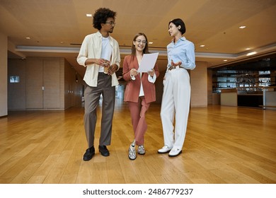 Three colleagues, diverse in ethnicity and background, stand in a modern office, engaging in conversation. - Powered by Shutterstock