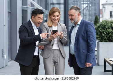 Three Colleagues Employees Watch Funny Videos Or Look At Photos, Pictures Memories Looking At Smartphone Screen Using Mobile Phone. Group Of Business People Office Workers Having Fun On Breaks