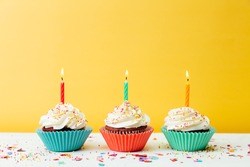 Three Colorful  Birthday Cupcakes With Candles And Confetti On A Yellow Background