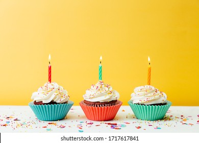 Three Colorful  Birthday Cupcakes With Candles And Confetti On A Yellow Background