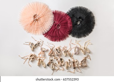Three colorful tutus and many beige pointe shoes on the light floor in the studio. Tutus are peach, burgundy and black colored. Top view photo. Horizontal.