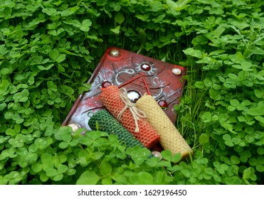 Three Colorful Wax Candles And Closed Magic Book In Clover Grass. Mystic Background With Esoteric, Divination And Gothic Objects, Wiccan And Pagan Concept.