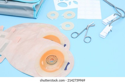 Three colostomy bags with medical scissors, a stencil of different diameters, a sticky circle, a bag and clothespins on a rope lie on a soft blue background, close-up side view.