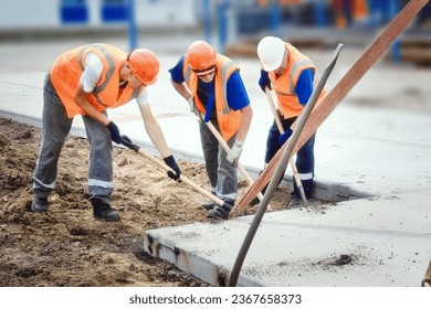 Three construction workers in vests and hard hats lay concrete slab at construction site. Team of workers with shovels lays slab on summer day. Authentic workflow.
