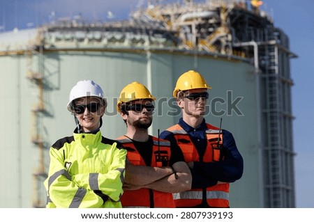Three construction workers wearing safety gear at an industrial site with large storage tanks visible in the background