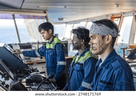 Three crew members operating navigation and maneuvering consoles on the bridge of a commercial cargo vessel during sea passage.
