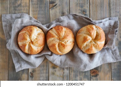 Three crusty round bread rolls, known as Kaiser or Vienna rolls in a row on linen towel on rustic wooden background, flat lay
