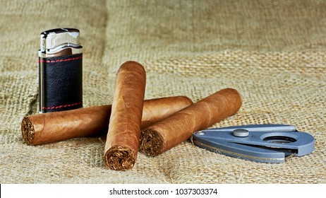 Three Cuban Cigars With A Lighter And A Cutter On A Jute Background