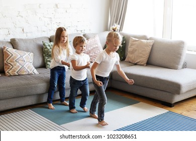 Three Cute Children Wearing Similar White Tops And Jeans Standing Barefooted On Carpet While Dancing Or Doing Conga At Home Or Kindergarten. Emotional Children Having Fun Indoors, Playing Games