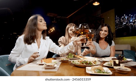 Three cute girlfriends having a rest sitting at a table and drinking wine merrily talking. Girls clink glasses with wine glasses. - Powered by Shutterstock