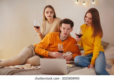 Three cute happy funny friends having fun together, arranging home cinema evening, watching comedy on laptop, laughing out loudly, sitting on bed with cheese plate and glasses of red wine