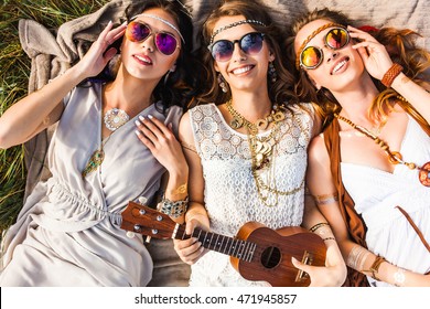Three cute hippie girl lying on the plaid outdoors, best friends having fun and laughing, play ukulele, sunglasses, feathers in their hair, bracelets, flash tattoo, indie, Bohemia, boho style top view