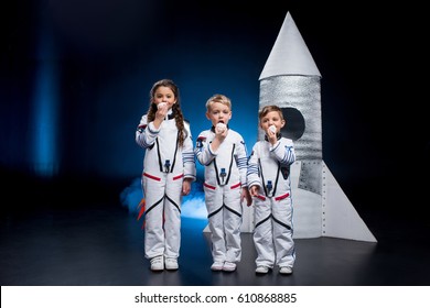 Three Cute Kids In Astronaut Costumes Eating Sweets Standing Near Rocket 