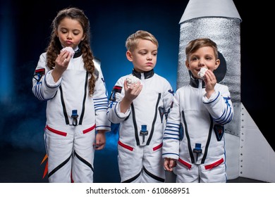 Three Cute Kids In Astronaut Costumes Eating Sweets Standing Near Rocket 