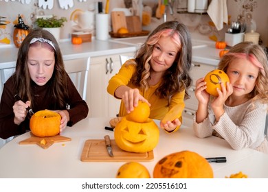 Three cute little sisters drawing   carving spooky faces pumpkins in light white kitchen at home  Halloween party preparation 