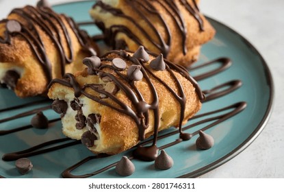 Three delectable pastries, generously filled with a creamy mixture and sweet cocoa morsels, are artfully drizzled with rich dark syrup and arranged on a vibrant teal plate.