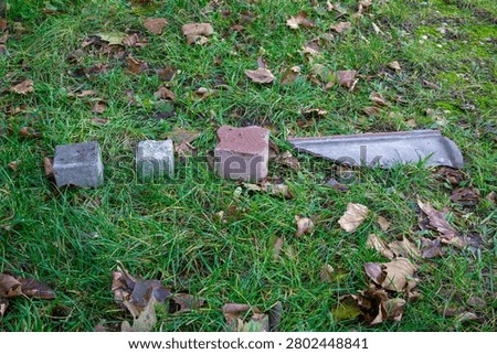 Three different blocks or cobblestones next to a piece of broken grey clay on green grass with brown leaves, group of stones in backyard, remnants of street repair or paving material