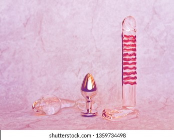 Three different sex toys (glass dildo, glass and metal butt plug) are on a light gray background with a stone texture. Image suitable for advertising sex shop