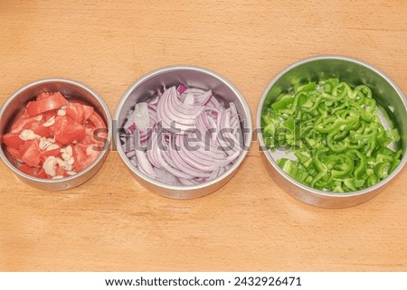 Three different sized circular metallic plates set from small to large, filled with raw tomatoes and garlics, slices onions and pieces of sliced green pepper. Separated and set on a wooden table top.