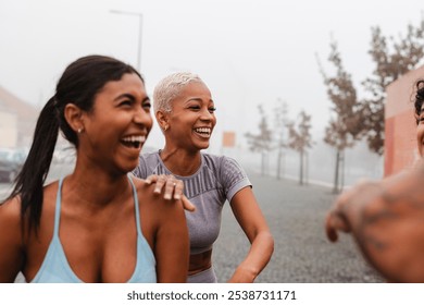 Three diverse female athletes are sharing a laugh together after a workout session in a city setting, demonstrating camaraderie and the joy of exercising - Powered by Shutterstock