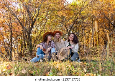 Three diverse female friends in cowboy hats are sitting on a log in the autumn forest. Female friendship. Outdoors. Cheerful person.