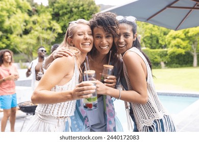 Three diverse female friends are embracing and smiling at an outdoor pool party. Diverse friends enjoy a sunny day together, with a young African American man visible in the background. - Powered by Shutterstock