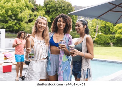 Three diverse female friends enjoy a poolside gathering, holding drinks and smiling warmly. A diverse group, they exhibit a relaxed, summery style in an outdoor social setting. - Powered by Shutterstock