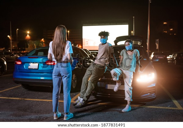 Three diverse friends wearing protective\
masks, standing by the car parked in front of a big screen. Young\
guys watching a movie in open air cinema. Safe entertainment during\
coronavirus quarantine