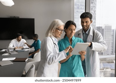 Three diverse general practitioners in uniforms working together, holding tablet device reviewing patient data, discussing medical case, sharing knowledge, using modern technology in medical practice - Powered by Shutterstock