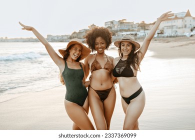 Three diverse girlfriends in swimwear having fun at the beach, women smiling cheerfully while embracing. Happy female friends enjoying their summer vacation - Powered by Shutterstock