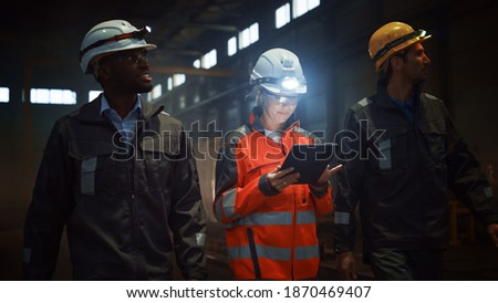 Three Diverse Multicultural Heavy Industry Engineers and Workers in Uniform Walk in Dark Steel Factory Using Flashlights on Their Hard Hats. Female Industrial Contractor is Using a Tablet Computer.