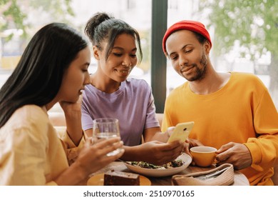 Three diverse, multinational friends using mobile phone sharing social media content while having lunch in a restaurant. Social media concept