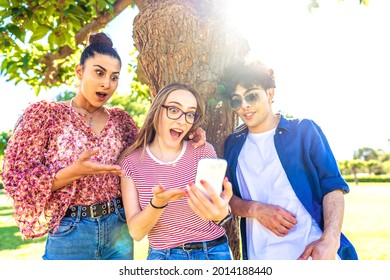 Three Diverse Students Making Surprised Faces Spreading Mouth And Eyes Pointing And Looking Smartphone Spending Time In Nature Of City Park. Power Of New Wifi Technology Addicting All Ages People