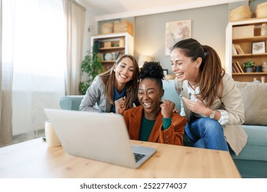 Three diverse women engage with a laptop, displaying excitement in a well-lit cozy living room, reflecting a moment of joy in a casual domestic setting. - Powered by Shutterstock