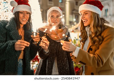 Three diverse women in Santa hats enjoy Christmas market festivities. Friends of different ethnicities and ages hold sparklers and drinks, laughing together amid twinkling lights. holiday spirit. - Powered by Shutterstock