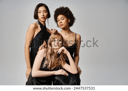 Three diverse women wearing black dresses strike a pose against a grey background in a studio setting.